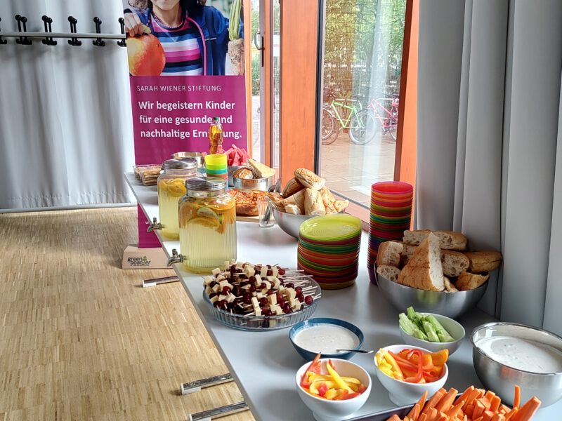 Ein aufgebautes Buffet im Vordergrund rechts. Dahinter das Banner der Sarah Wiener Stiftung